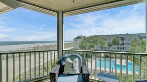 Un balcon avec un fauteuil en osier et une petite table, donnant sur une plage et une zone avec piscine.