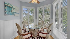 Un coin repas avec une table en verre entourée de chaises en osier, des murs recouverts de panneaux blancs et une vue sur les arbres à travers de grandes fenêtres.