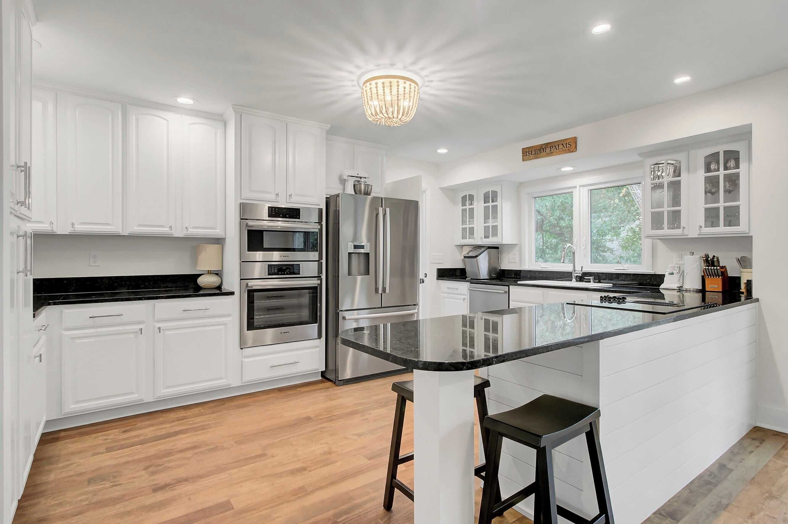 A modern kitchen with white cabinetry, stainless steel appliances, a large island with bar stools, and a chandelier.