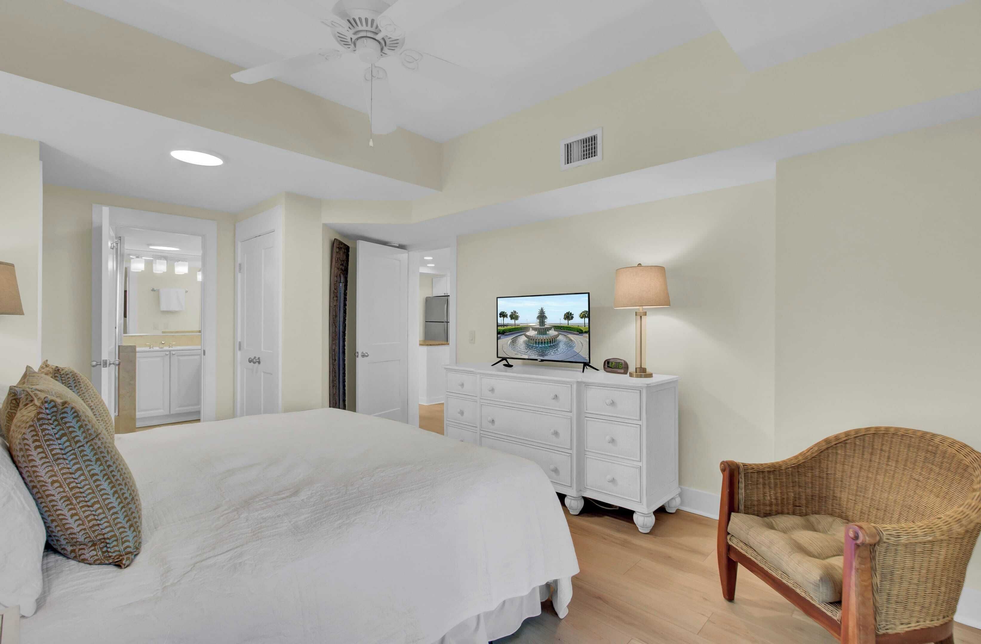 A bedroom with a large bed, a dresser, a television, and a wicker chair.