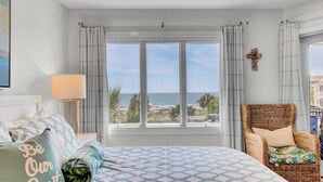 A bedroom with a large bed, a wicker chair, a view of the beach, and a cross on the wall.