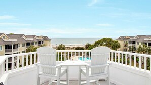 A balcony with two white Adirondack chairs, a small table, and a view of the beach and ocean.