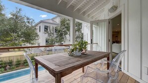 A wooden table with chairs on a covered patio overlooking a swimming pool, with a view of a house and trees in the background.