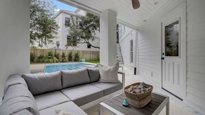 A covered outdoor seating area with a pool, a white building in the background, and a wooden fence.