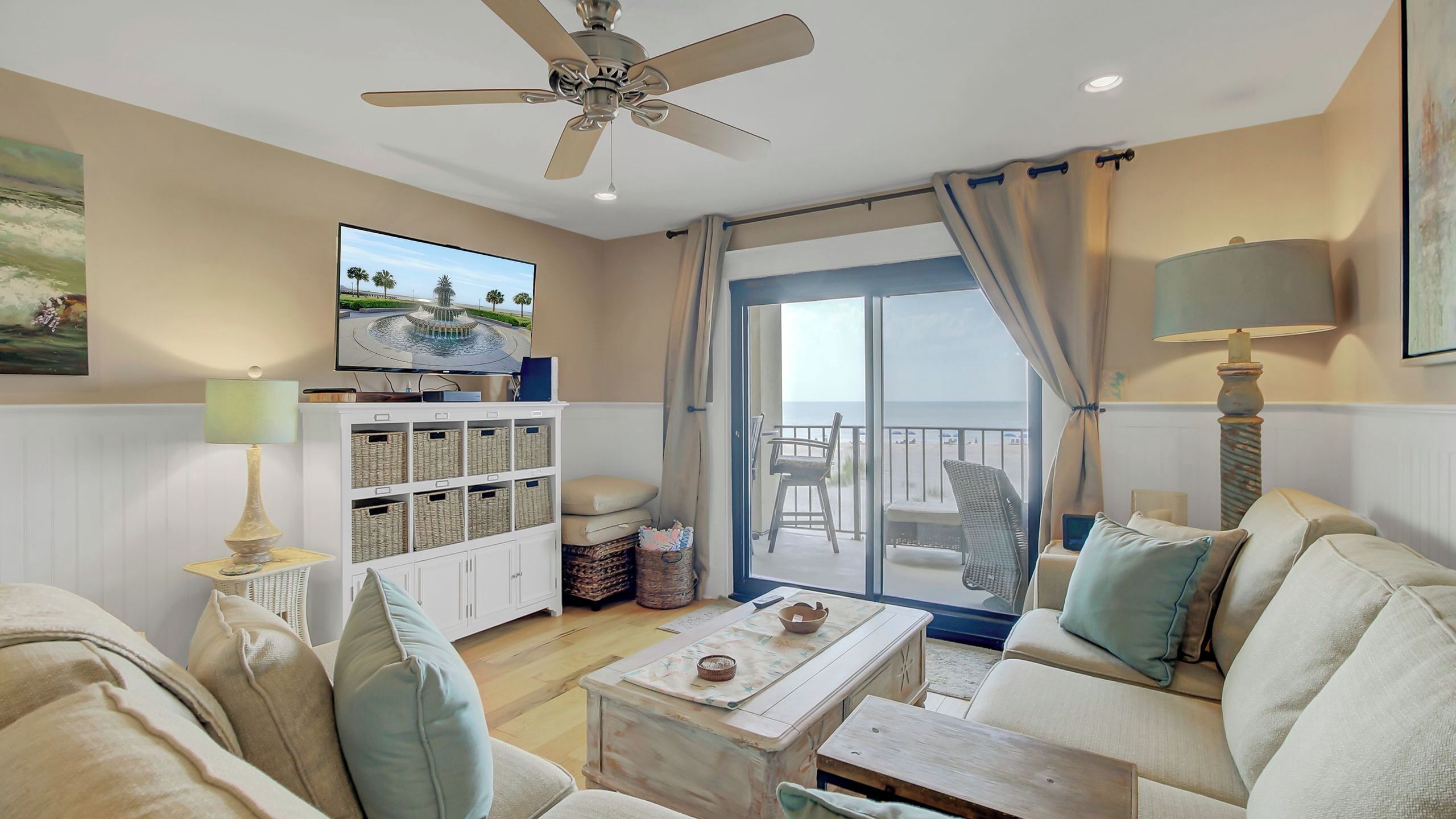 A living room with a ceiling fan, a television, a sofa, a coffee table, and a view of the sea through a sliding glass door.