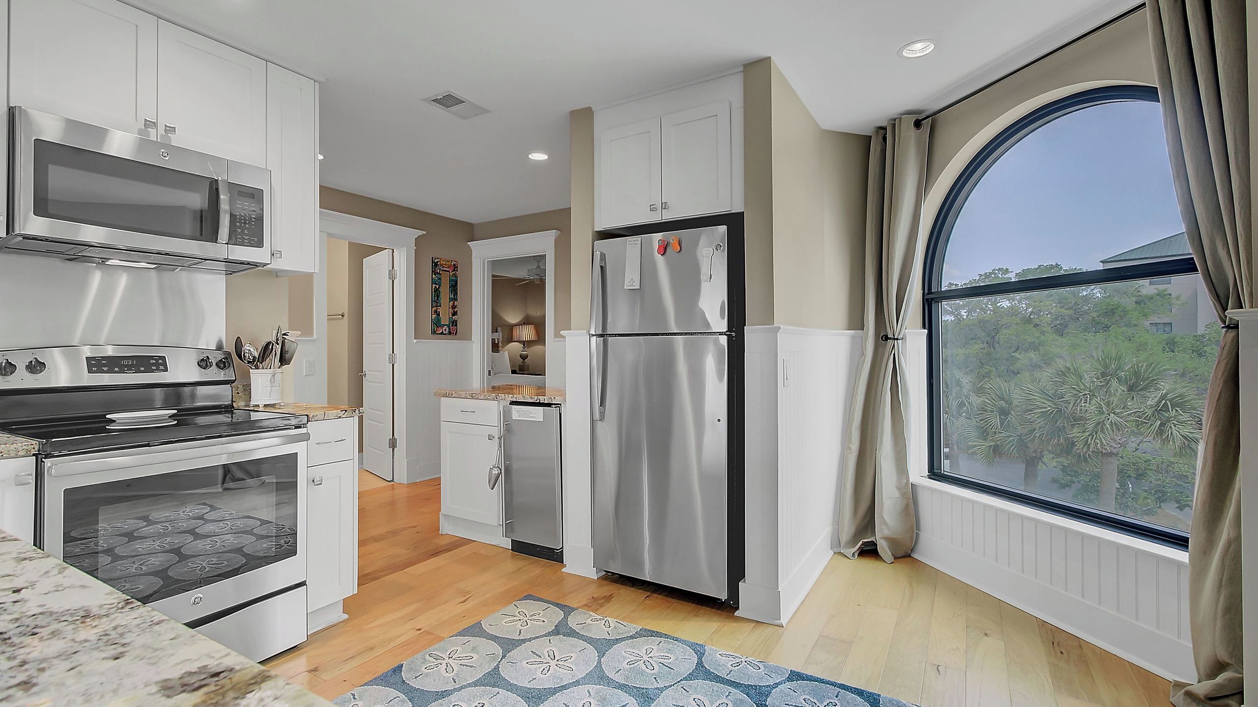 A modern kitchen with stainless steel appliances, a large window with curtains, and a view of greenery outside.