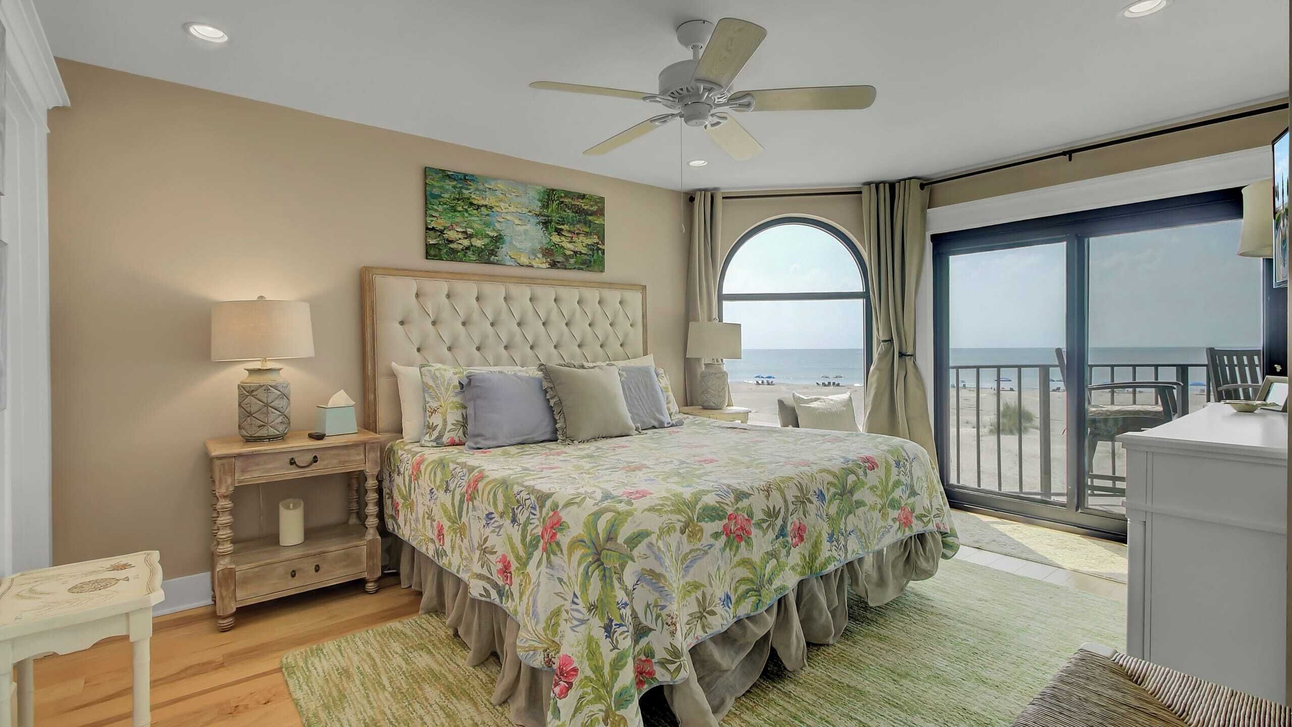 A bedroom with a large bed, a ceiling fan, a painting, a nightstand, a desk, and a view of the beach.