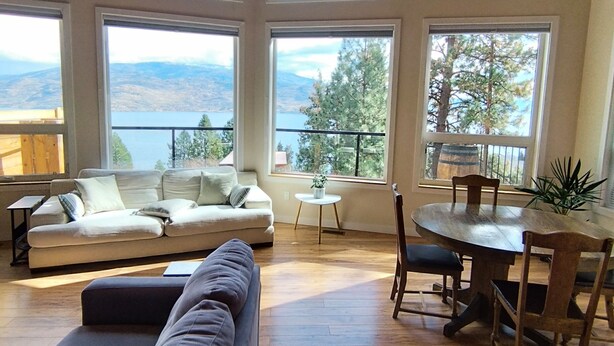 Ein geräumiges Wohnzimmer mit hohen Decken, großen Fenstern mit Blick auf Berge und Bäume, einem Holzesstisch mit Stühlen und einem grauen Sofa.
