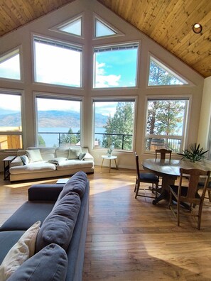 Un salon spacieux avec un plafond haut, de grandes fenêtres offrant une vue sur les montagnes et les arbres, une table à manger en bois avec des chaises, et un canapé gris.