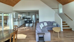 Ein modernes Interieur mit Holzbalkendecke, einer gebogenen grauen Couch, einem Esstisch mit Stühlen und einer Küche im Hintergrund.