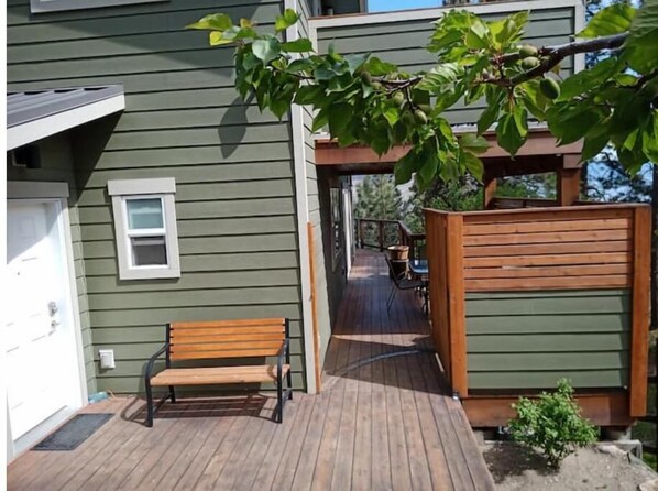 Una terraza de madera con un banco, una pérgola con plantas trepadoras y vista a una casa con garaje.