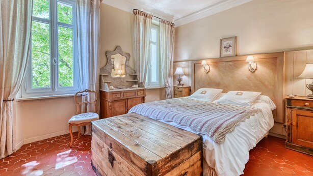 Une chambre à coucher avec une tête de lit en bois, un grand lit avec du linge de lit blanc, une commode en bois, une coiffeuse avec un miroir et une fenêtre avec des rideaux.
