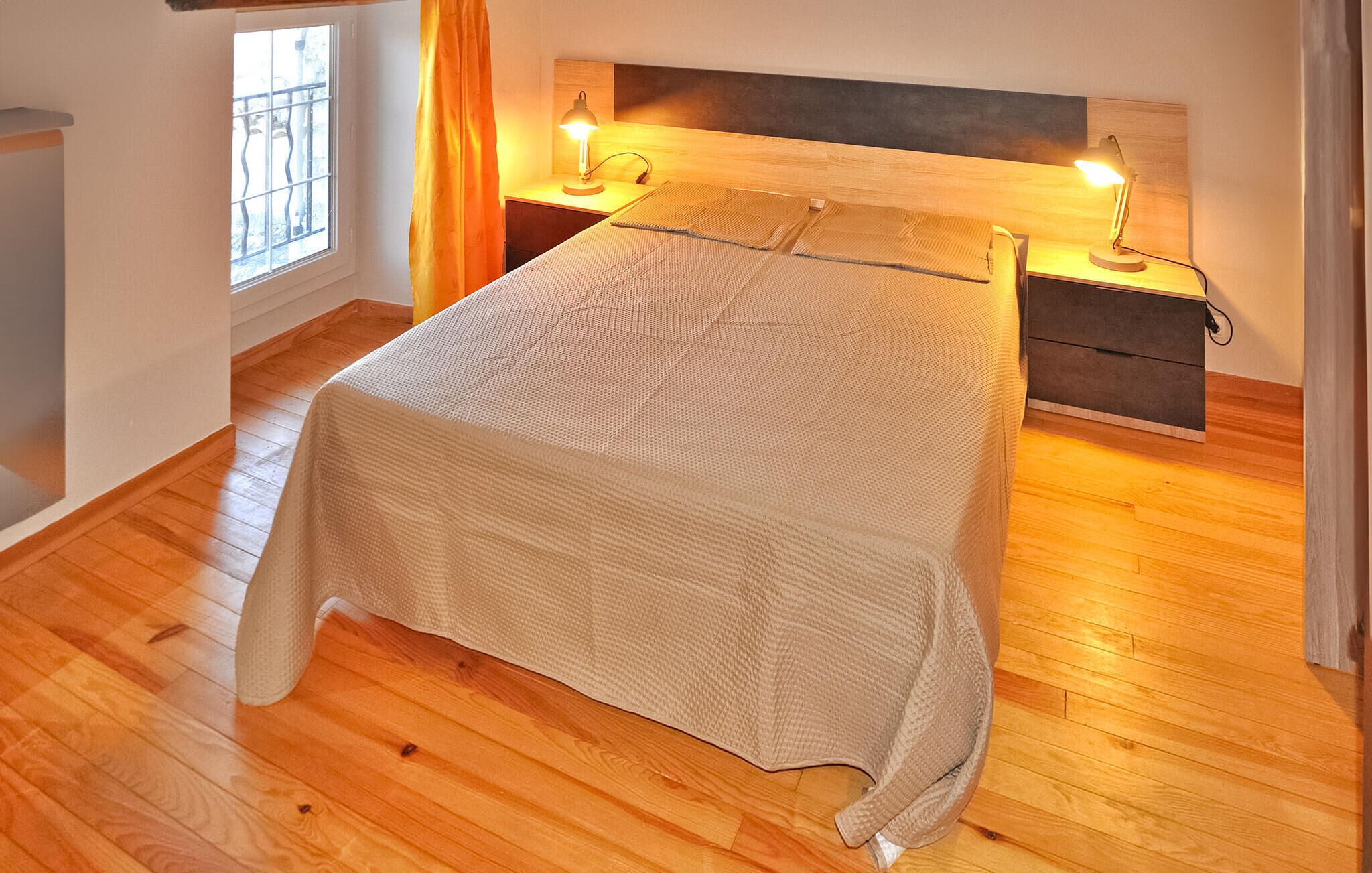 A bedroom with a wooden floor, a bed with a beige blanket, two bedside tables with lamps, and a window with a view of the outside.