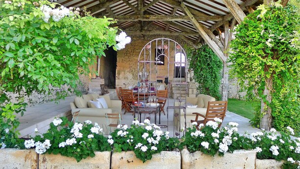 An outdoor seating area with stone walls, a wooden pergola, and white flowers.