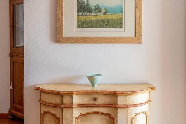 A vintage-style dresser with a bowl on top, a framed landscape painting above it, and a wooden cabinet to the left.