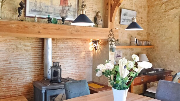 L’intérieur est de style rustique, avec des murs en briques apparentes, des poutres en bois et un poêle d’époque. On y trouve deux lampes suspendues, un vase rempli de fleurs et une table en bois avec des chaises.