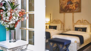 A hotel room with two beds, a large painting on the wall, a vase of flowers on a table, and a view of the outside. - The Royal Hermitage (Jaipur)