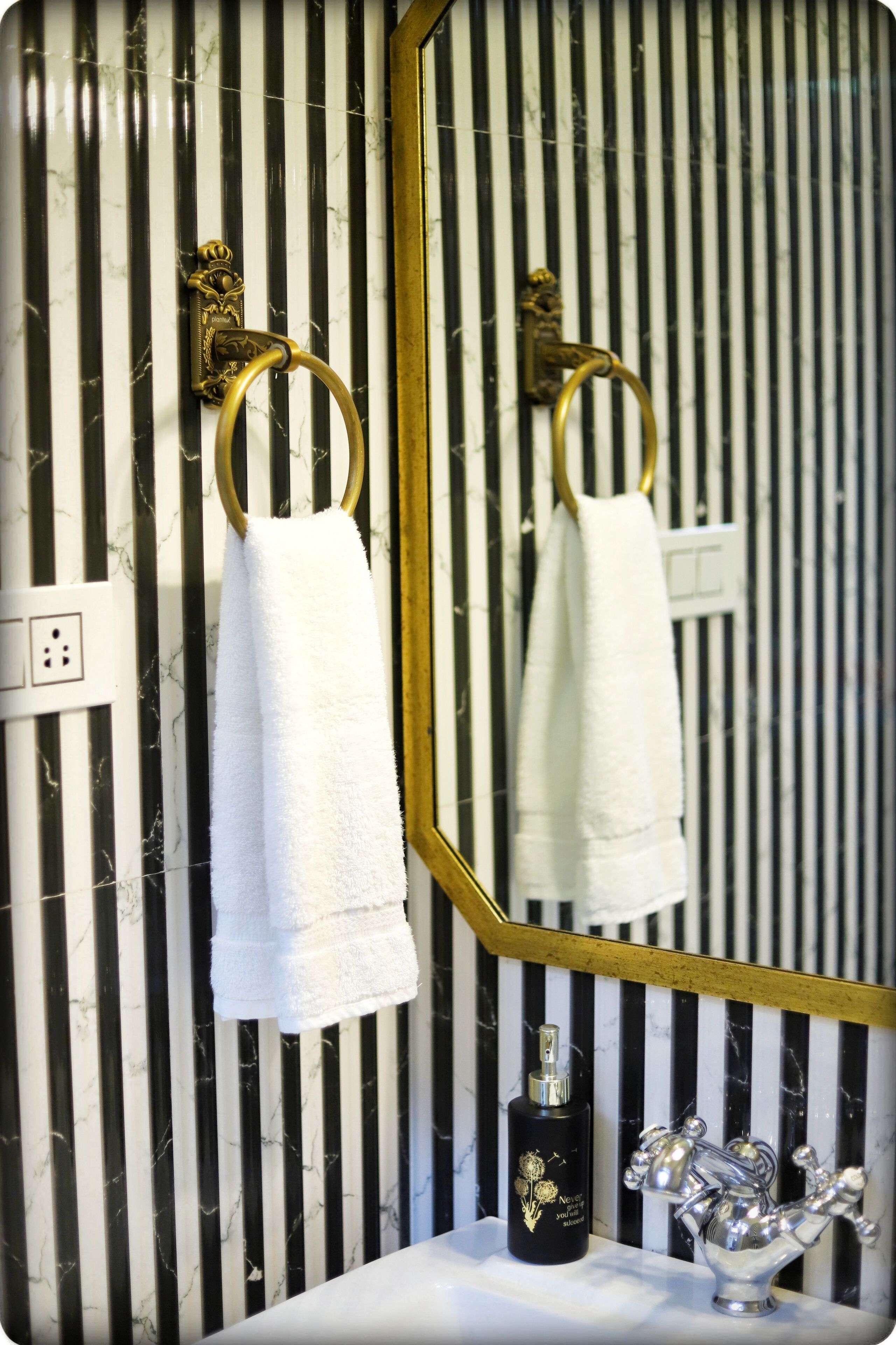 A bathroom with a striped marble wall, a gold-framed mirror, a white towel hanging on a gold towel ring, a black bottle with gold detailing, and a silver faucet.