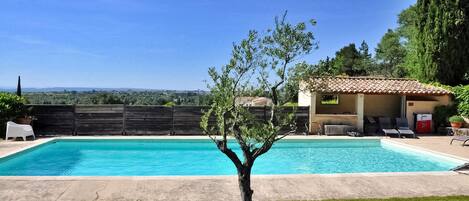 Una piscina con cubierta de madera, un árbol pequeño y un edificio con tejado de tejas al fondo.