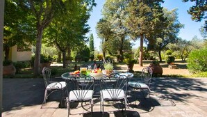Une terrasse avec une table ronde et des chaises, entourée d’arbres et de plantes en pot.