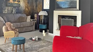 A living room with a grey sofa, a red armchair, a blue ottoman, and a teddy bear. There is a fireplace with a white mantel and a framed picture above it.