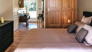 A bedroom with a large bed, a wooden wardrobe, a small table with a chair, and a view of the outdoors through a glass door.