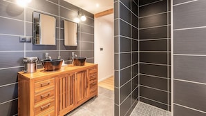 A modern bathroom with dark tiled walls, two sinks with copper bowls, wooden cabinetry, and a shower area with a dark tiled shower enclosure.