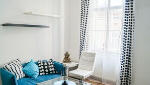Un salón con un sofá azul, una mesa de centro de cristal, un sillón blanco y una ventana con cortinas a lunares.