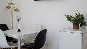 Una habitación con una mesa blanca, sillas negras, una planta pequeña en maceta, un armario blanco y una lámpara de pie.