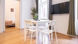 A room with a white table and chairs, a potted plant, a TV mounted on the wall, and a bed in the background.