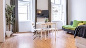 A modern living room with a flat-screen TV, a dining table with chairs, a green sofa, and a potted plant.