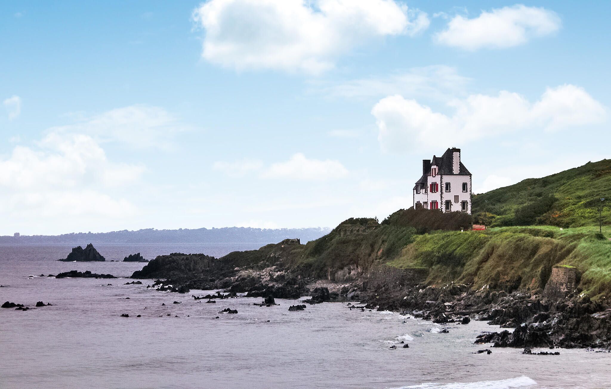 Ein Küstenhaus mit weißer Fassade und roten Akzenten, auf einem grasbewachsenen Felsen mit Blick auf das Meer.