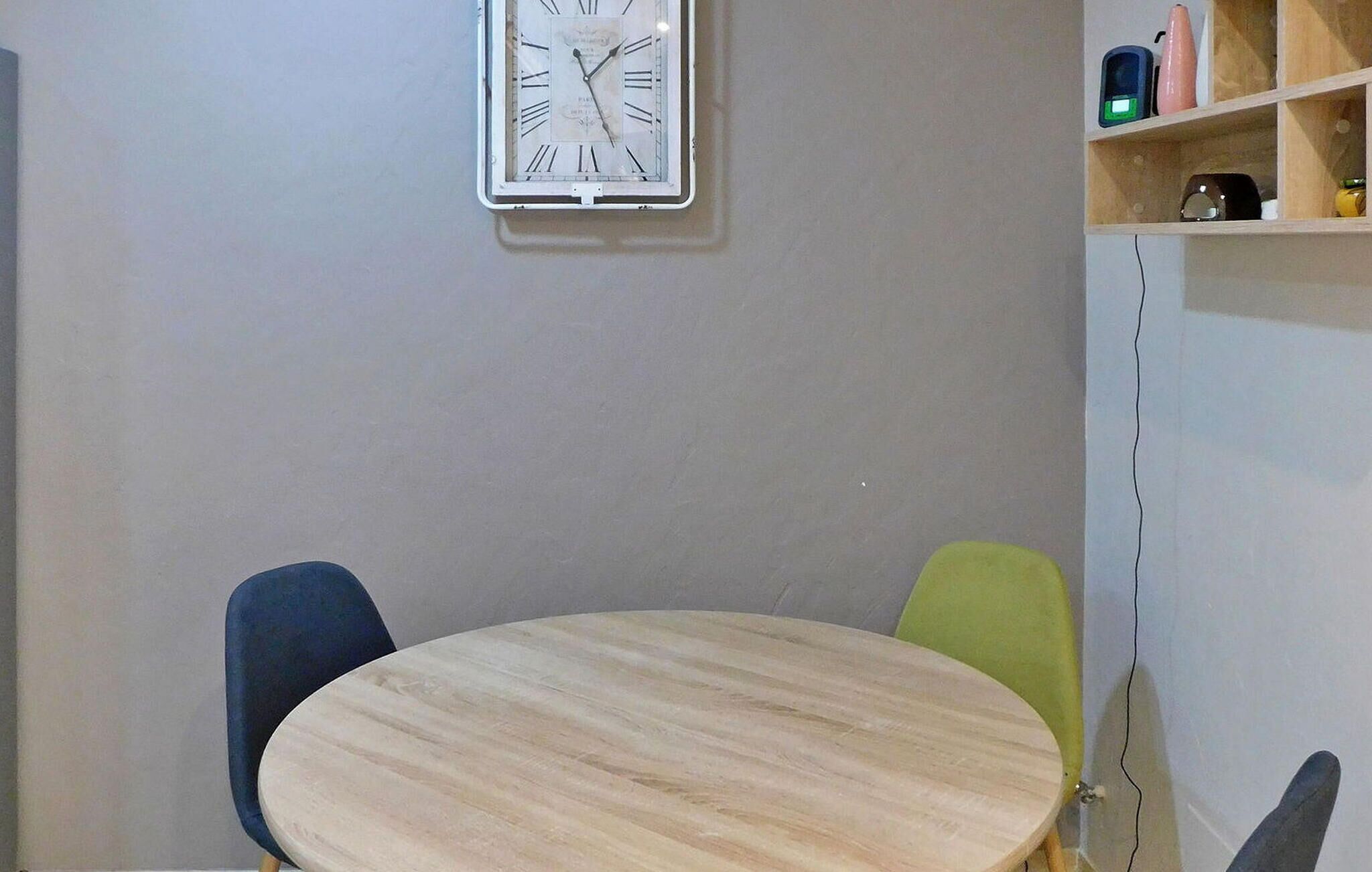 A room with a round wooden table, two chairs, a white clock on the wall, and a shelf with various items.