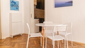 Une petite cuisine équipée d’un réfrigérateur, de placards et d’un évier. On y trouve une table blanche avec quatre chaises et un tableau accroché au mur.