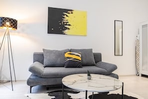 Uma sala de estar moderna com um sofá cinzento, uma mesa de centro redonda de mármore, um abajur e uma obra de arte na parede.
