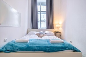 A hotel room with a bed, a window with curtains, a nightstand with a lamp, and a wall-mounted light.