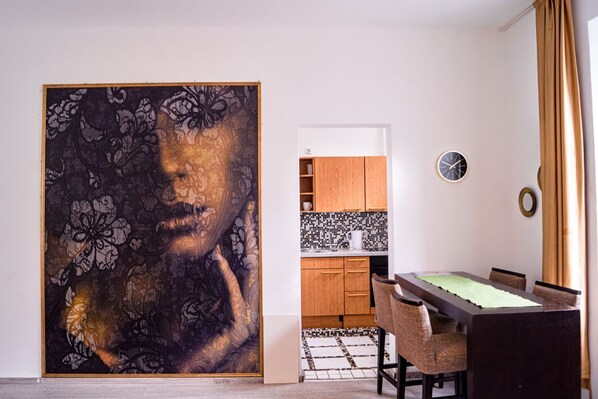 A dining area with a table and chairs, a kitchen in the background, and a large artwork on the wall.