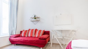Una habitación con un sofá rojo, un escritorio blanco, una pequeña estantería con plantas y una ventana con cortinas estampadas.