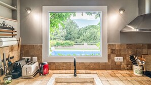Cuina amb una finestra gran que dona a un jardí i una piscina, equipada amb torradora, bullidor i diversos utensilis.