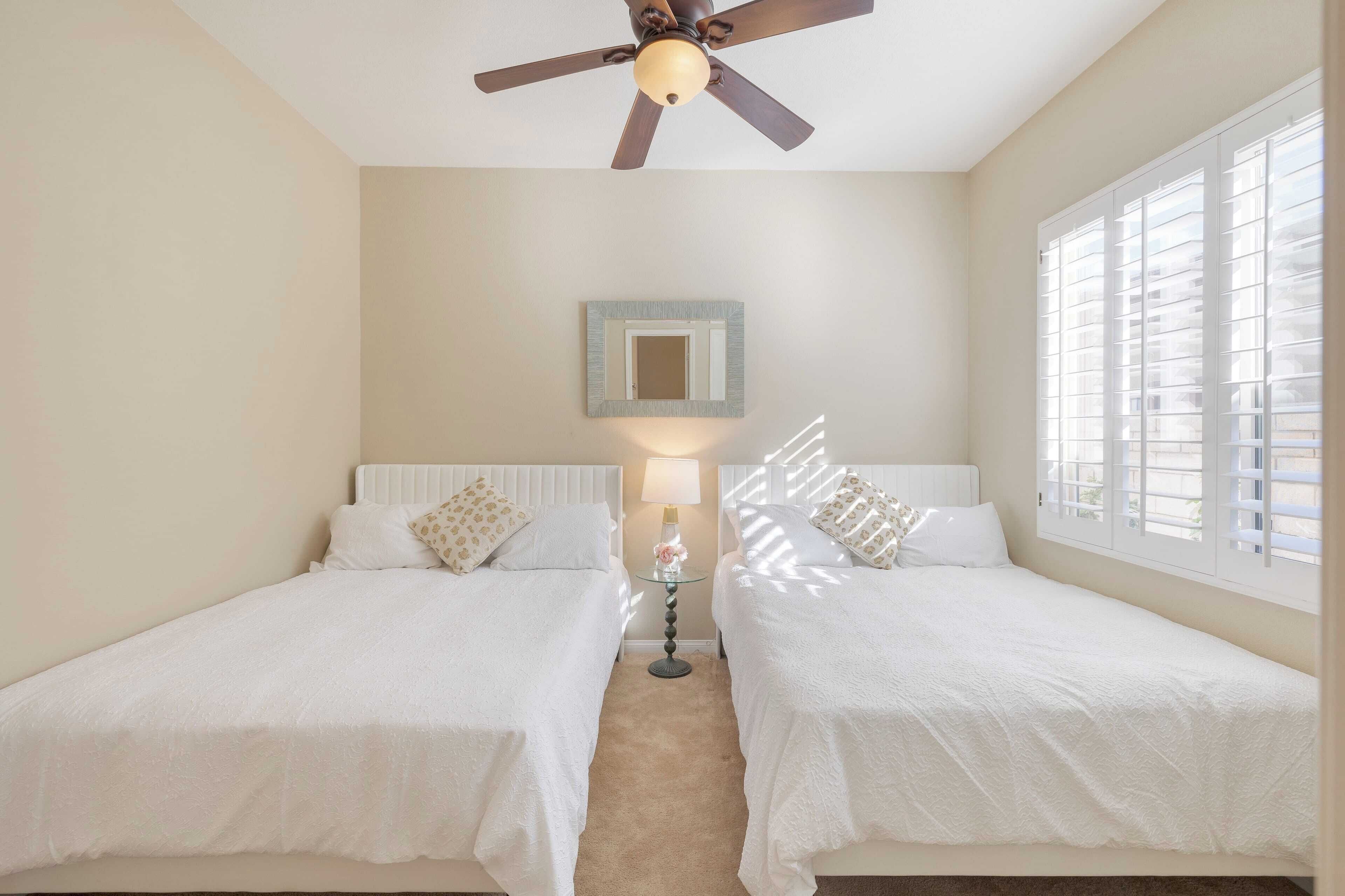 A hotel room with two single beds, a ceiling fan, a bedside table with a lamp, a mirror on the wall, and a window with blinds.