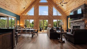 A spacious interior with a high ceiling, wooden walls, a dining area, a living room with a fireplace, and large windows offering views of the outdoors. - Luxe Lakefront Retreat w/ Pool, Hot Tub, Theater & Private Dock Sleeps 20+ (Sherrills Ford)