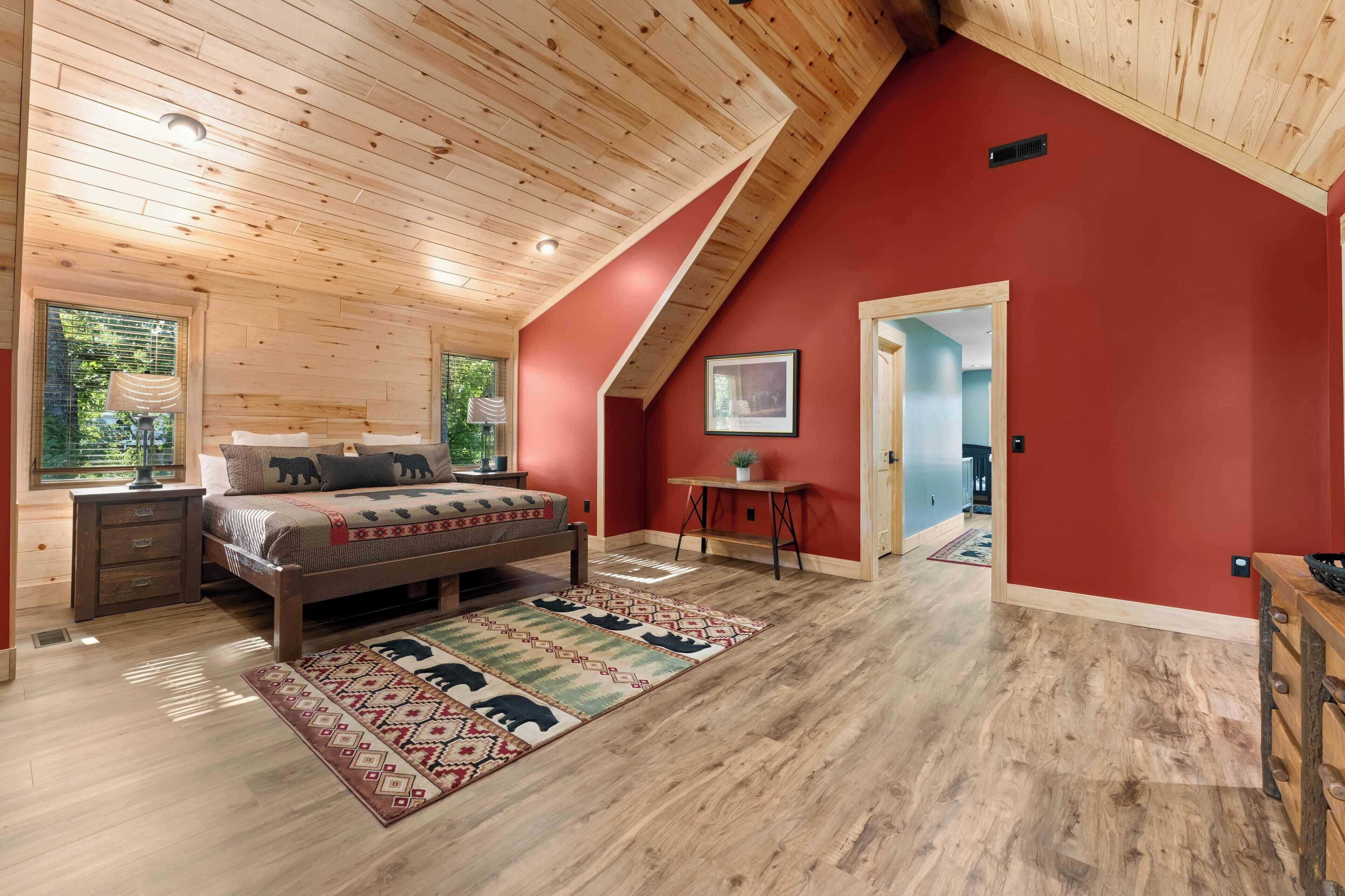 A bedroom with a wooden ceiling, a bed with a patterned bedspread, a small table, and a red accent wall.