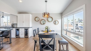 A modern kitchen with white cabinets, a dining area with a wooden table, and large windows offering a view of the neighborhood. - Ocean Breeze II by Avantstay Steps to the Beach! (Newport Beach)