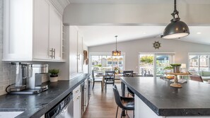 A modern kitchen with a central island, stainless steel appliances, and a large window overlooking a deck. - Ocean Breeze II by Avantstay Steps to the Beach! (Newport Beach)