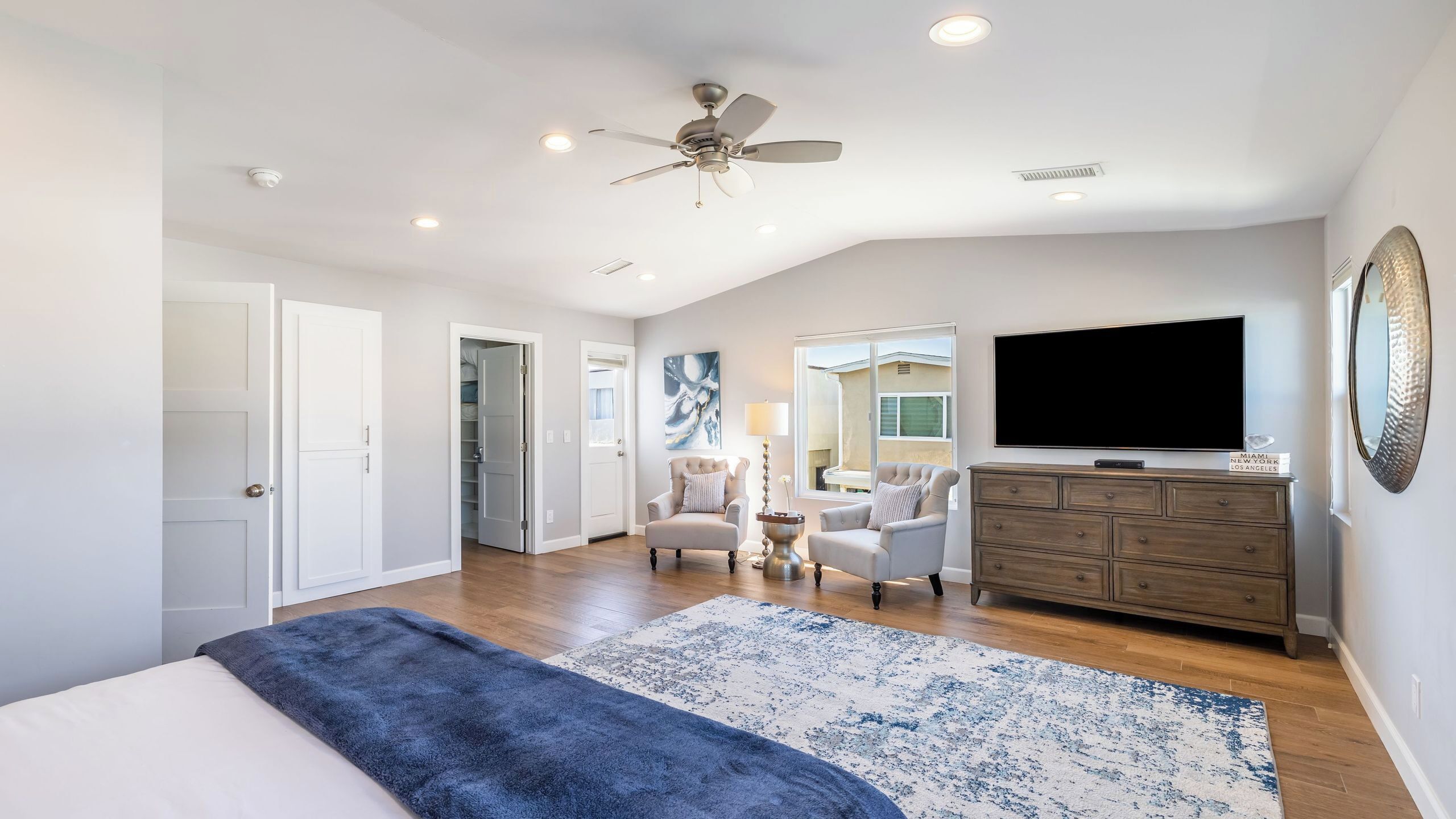 A bedroom with a bed, a ceiling fan, a TV, a dresser, and a view of the outside.