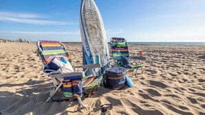 A beach scene with two striped beach chairs, a surfboard, and a bag labeled "AVANTIS" on the sand. - Ocean Breeze II by Avantstay Steps to the Beach! (Newport Beach)