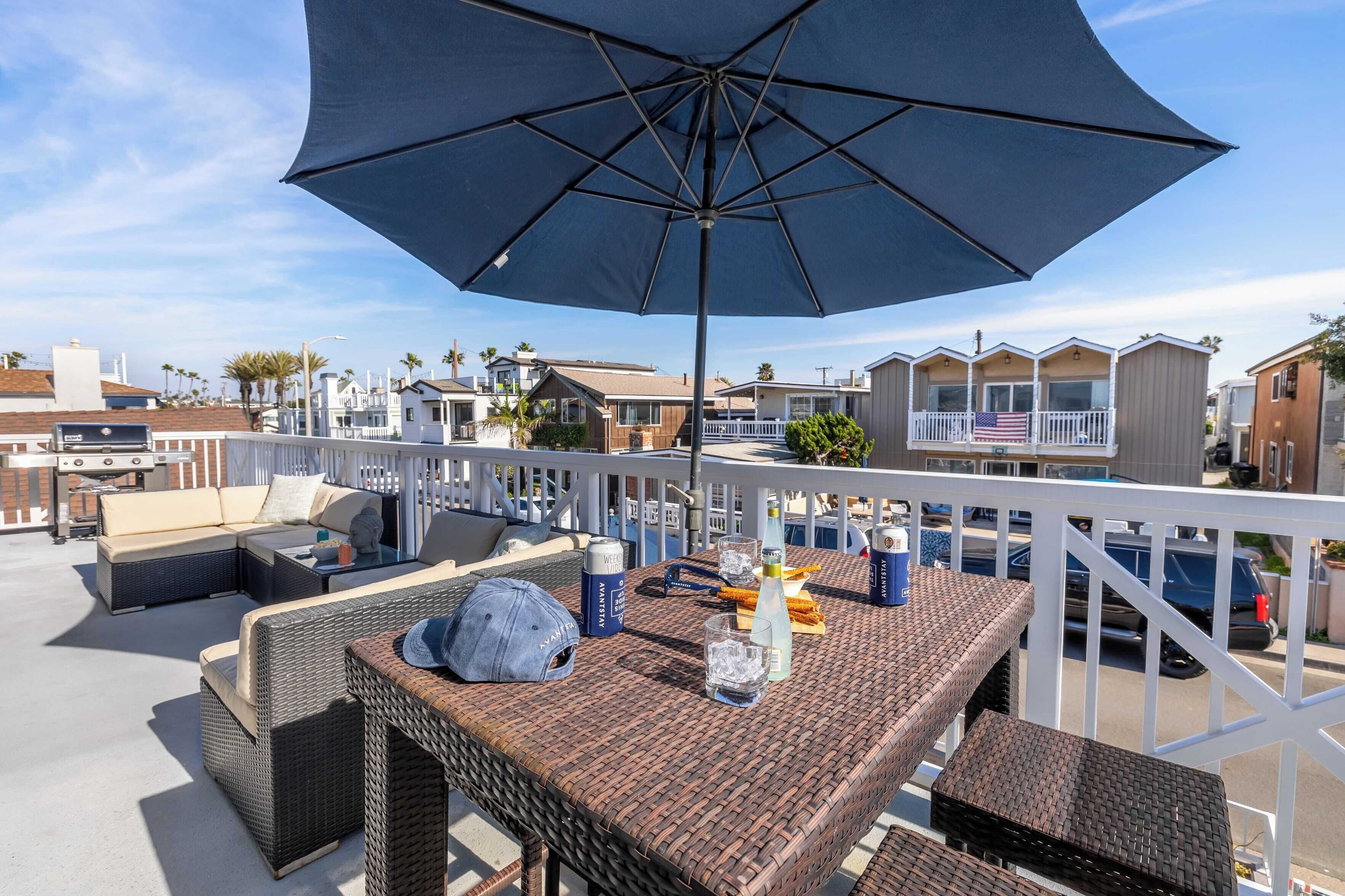 A rooftop patio with wicker furniture, a large umbrella, and a view of neighboring houses.