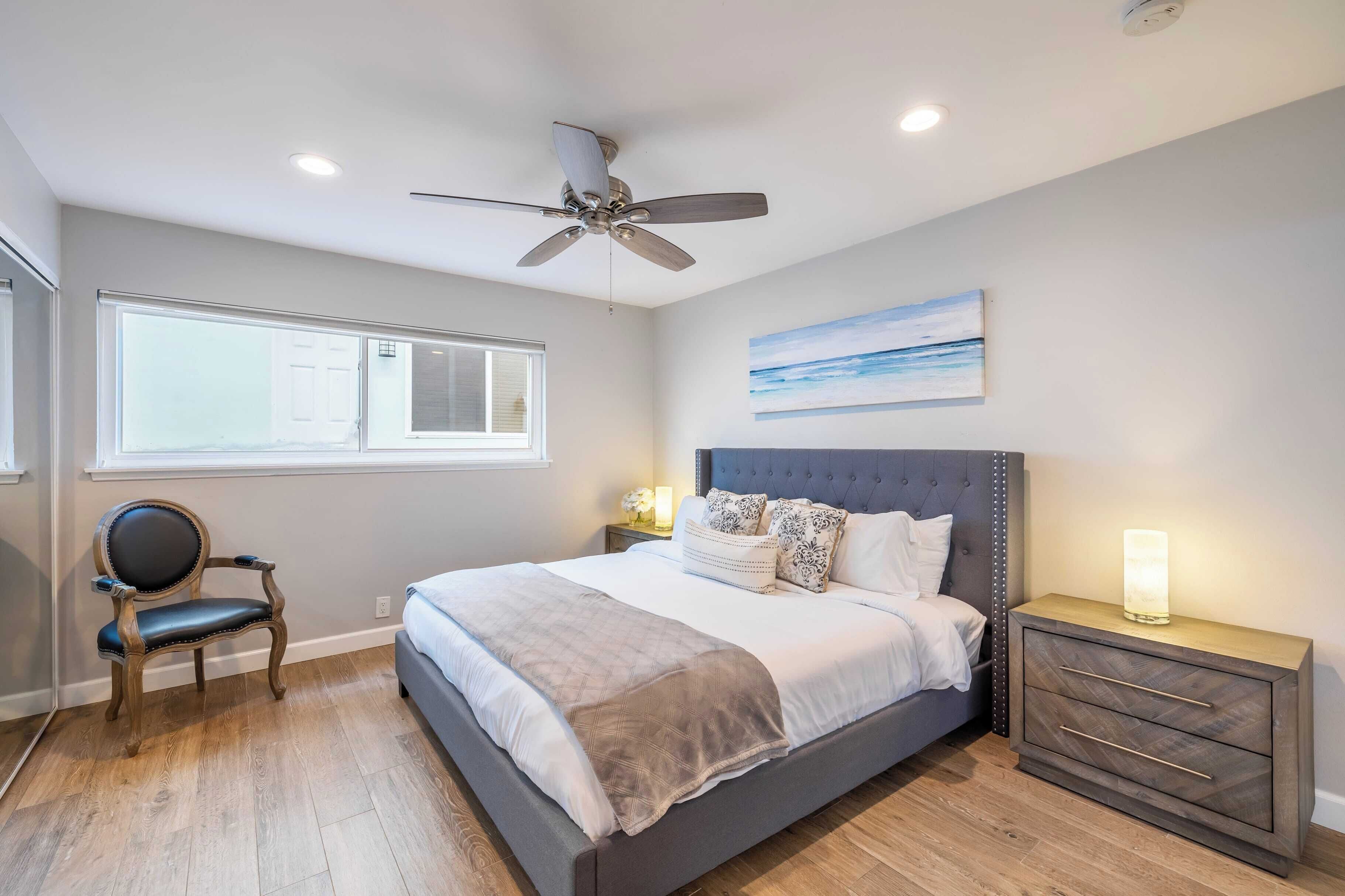 A bedroom with a bed, a nightstand, a chair, a ceiling fan, and a window.