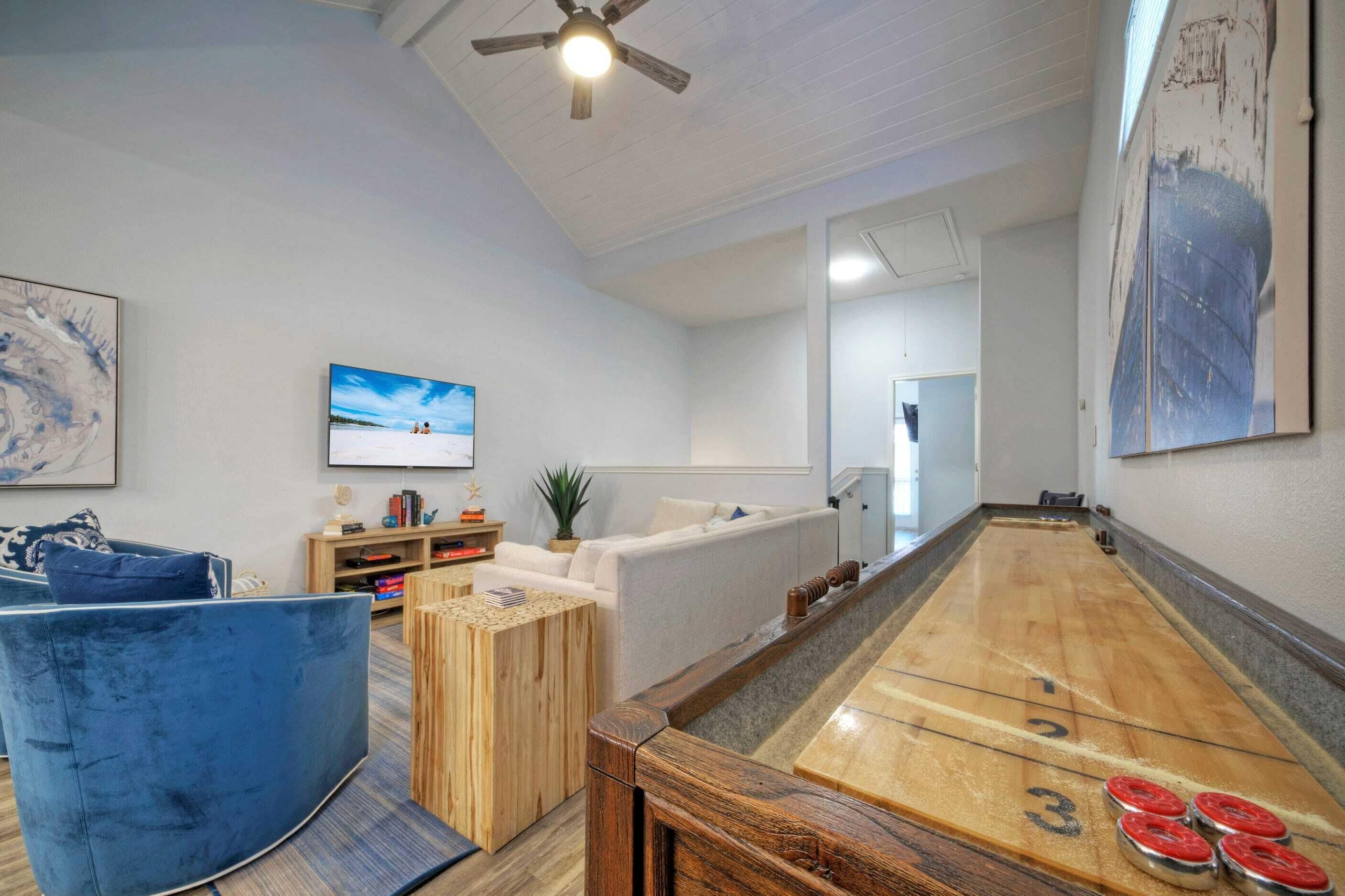 A room with a shuffleboard table, a blue sofa, a wooden coffee table, and a television on the wall.