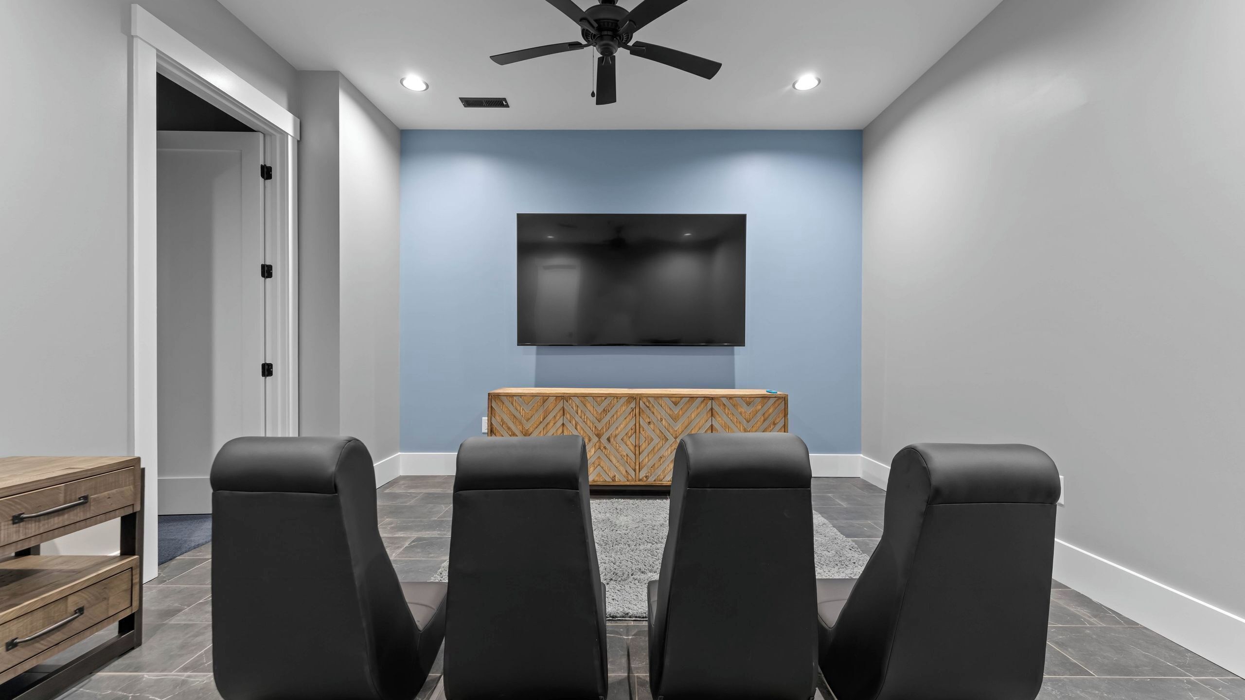 A modern home theater room with a flat-screen TV, ceiling fan, and black leather seating.